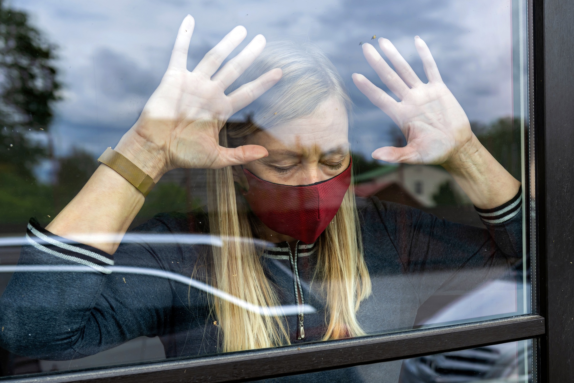 depresive woman in face mask looking out the window, staying home in quarantine, covid-19 lockdown